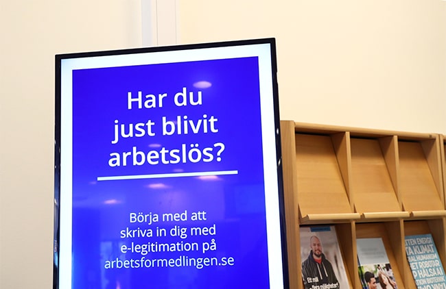 Blå arbetsförmedlingsskylt med text, med tidningsställ i bakgrunden.